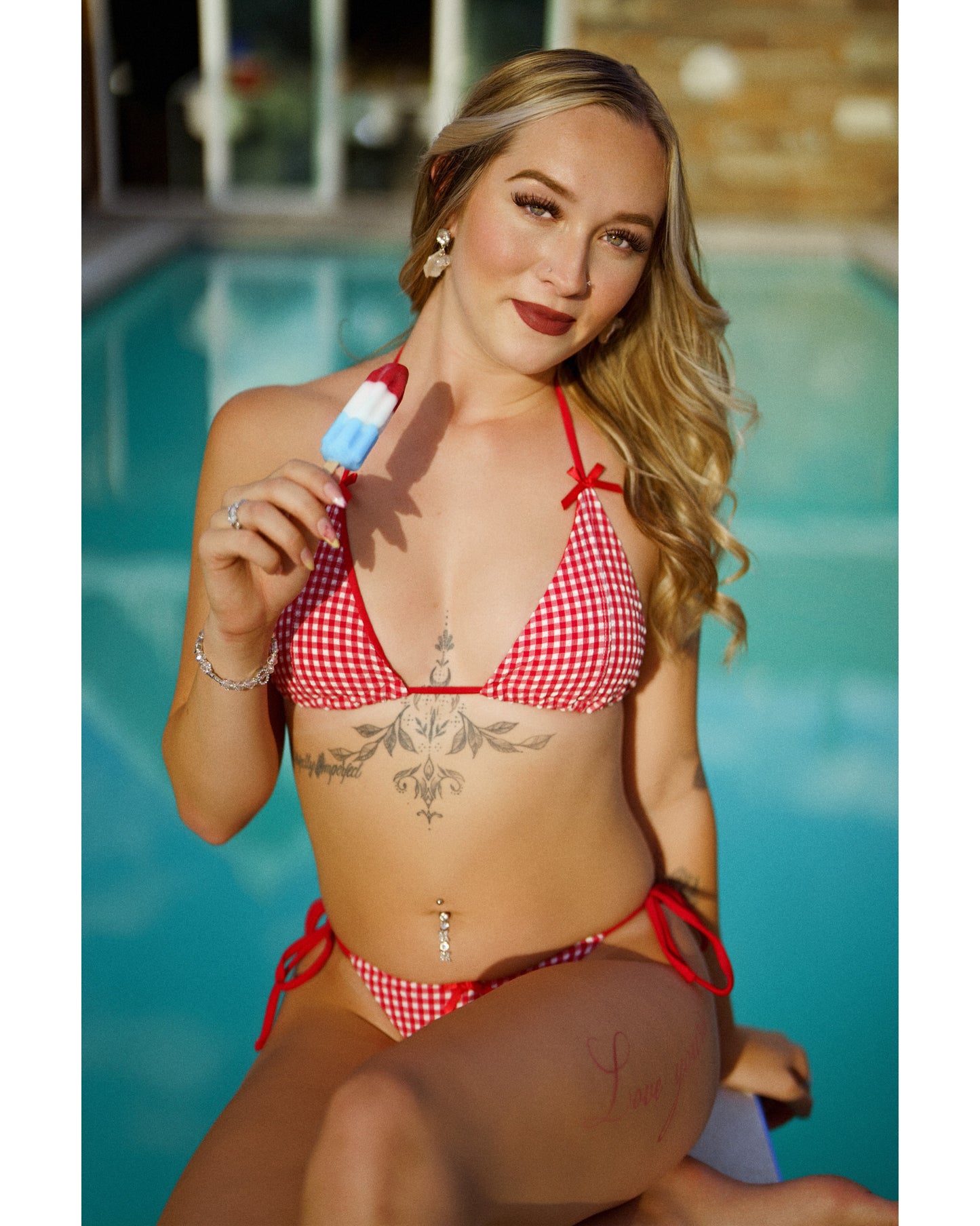 A woman wearing a red and white gingham string bikini sits by a swimming pool, holding a popsicle. 