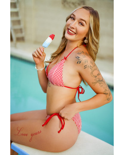 A woman wearing a red and white gingham string bikini sits on a swimming pool diving board, holding a red, white, and blue popsicle. The background shows a light-colored pool deck and water.