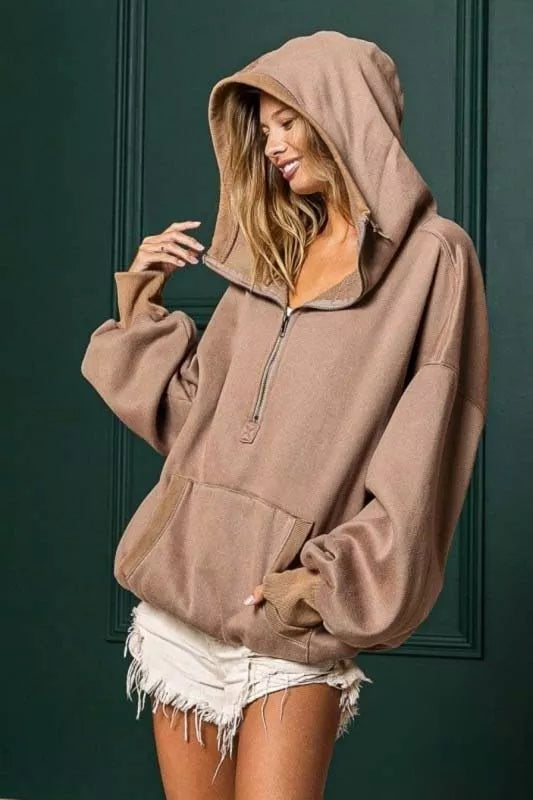 Woman wearing our mocha colored BiBi half zip fleece hoodie against a green wall, looking down and to the side while smiling.